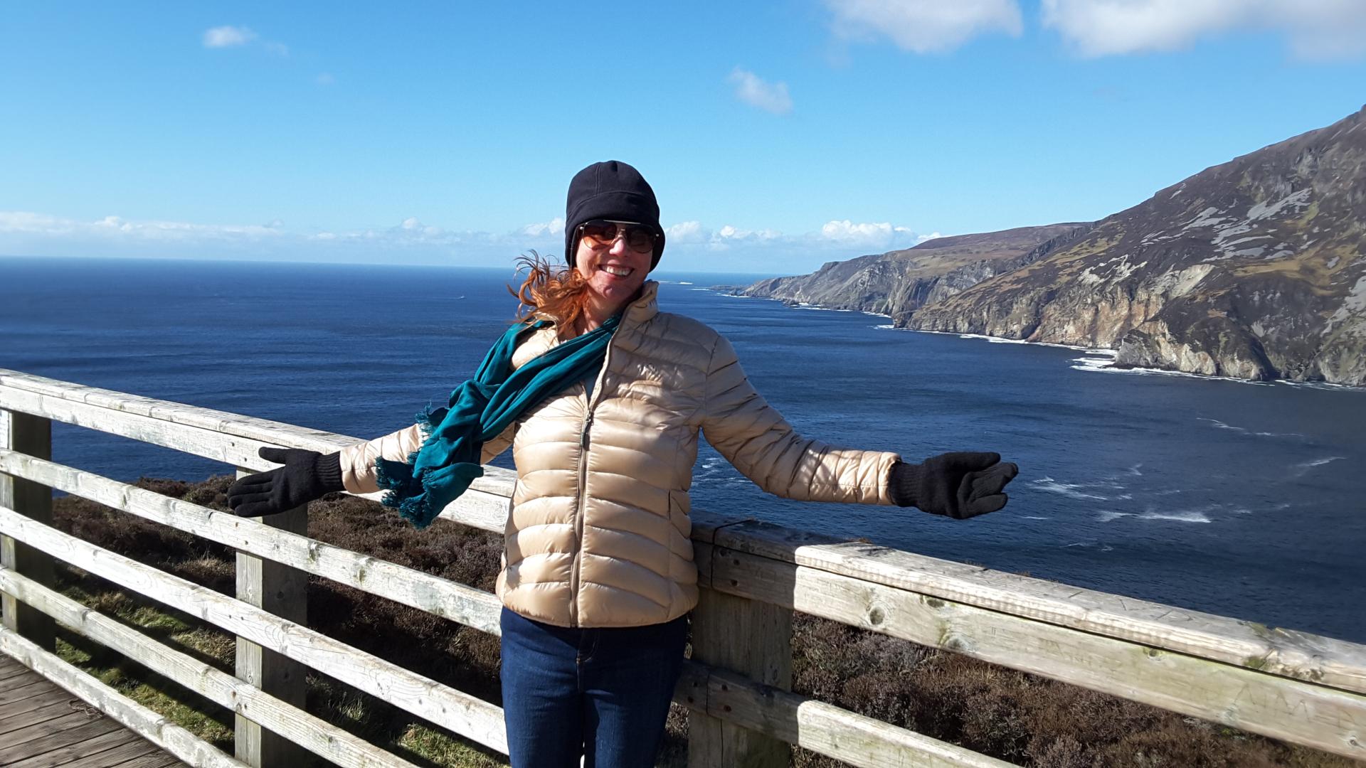 A smiling woman, arms open, standing with the ocean and spectacular cliffs behind her